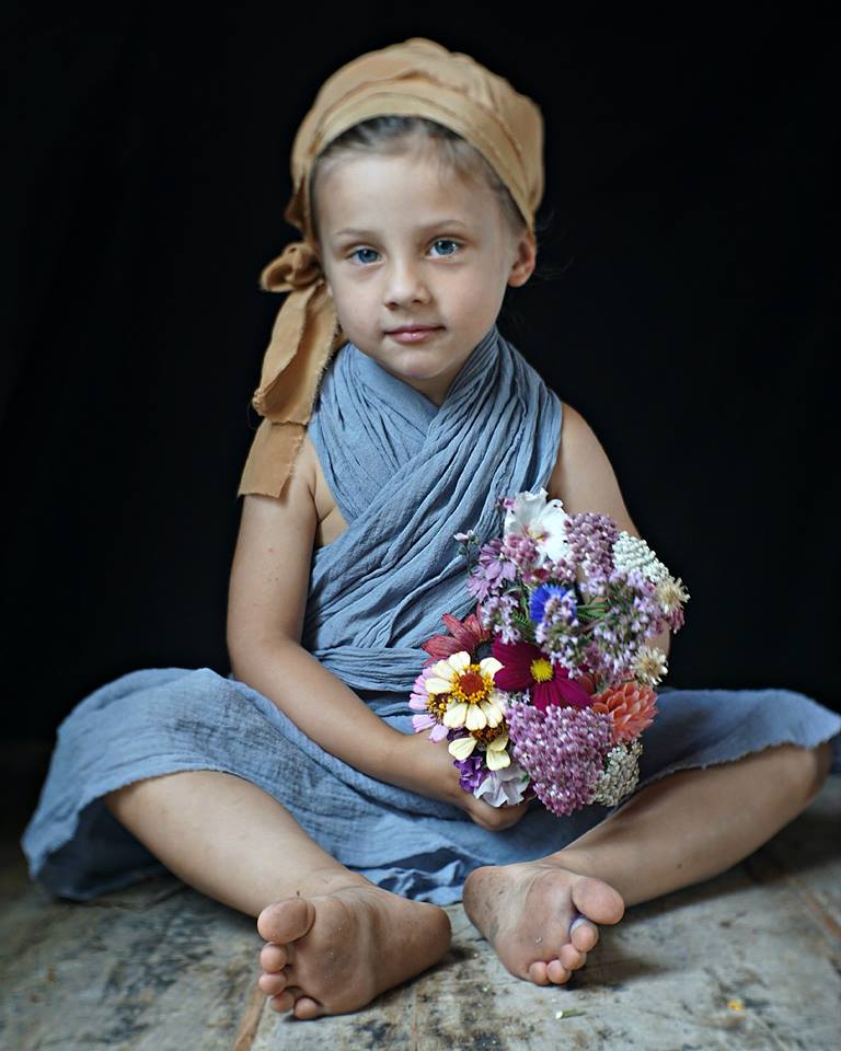 A child in a brown head covering sits while holding a fresh flower bouquet by Emily Avenson