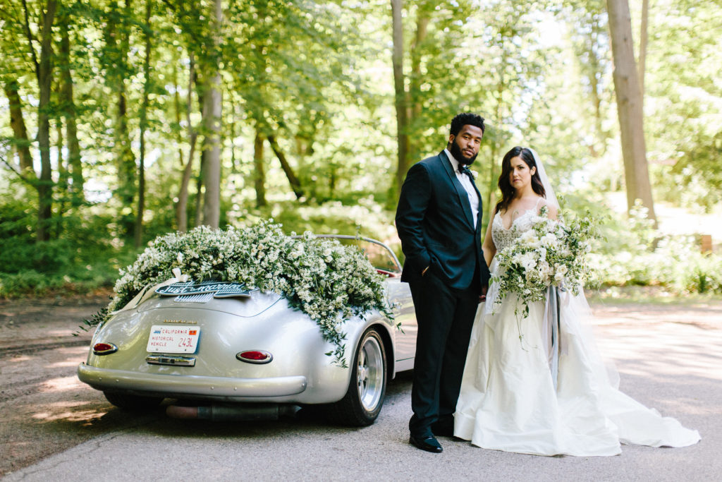 A bride and groom next to a classic silver Porsche outfitted with luxury floral arrangements
