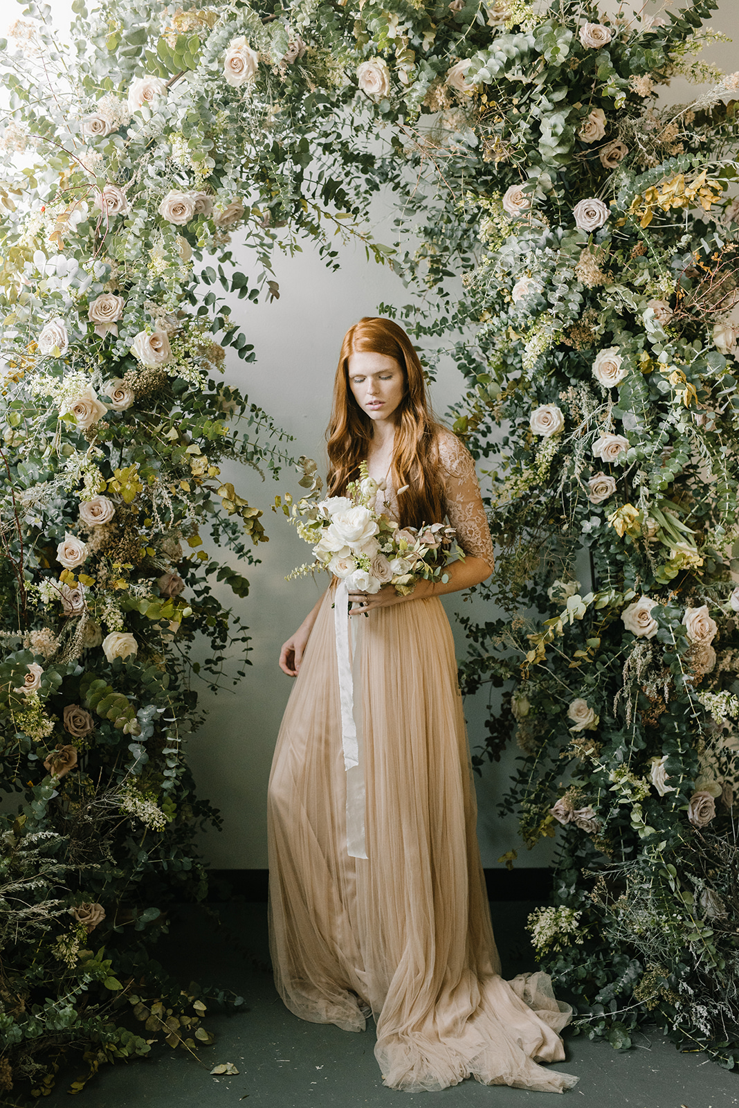 A model bride stands under a thick floral wreath with white roses holding a white bridal bouquet
