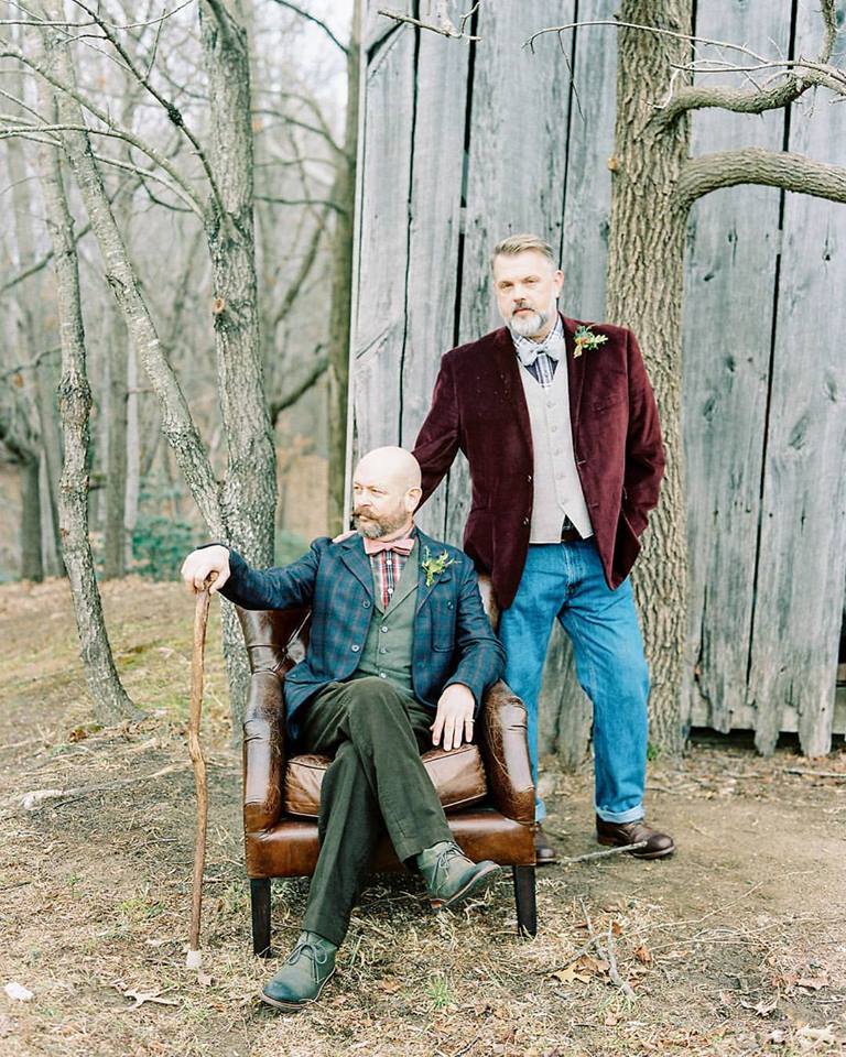 Brian Watson stands next to his husband Gene who is sitting in a brown leather chair in the woods