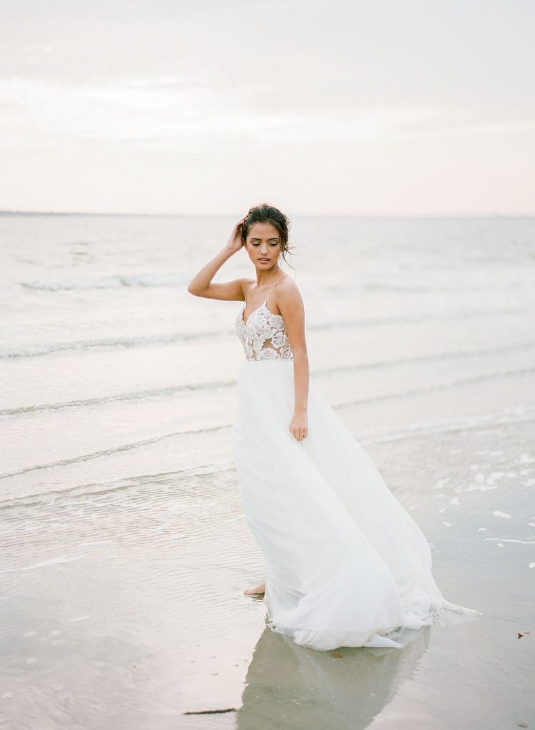 A bridal photo shoot model standing by the water on the beach in her a wedding dress