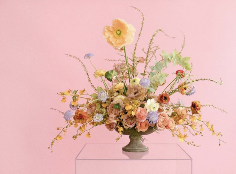 A floral arrangement sits on a clear plastic stand in front of a pink backdrop
