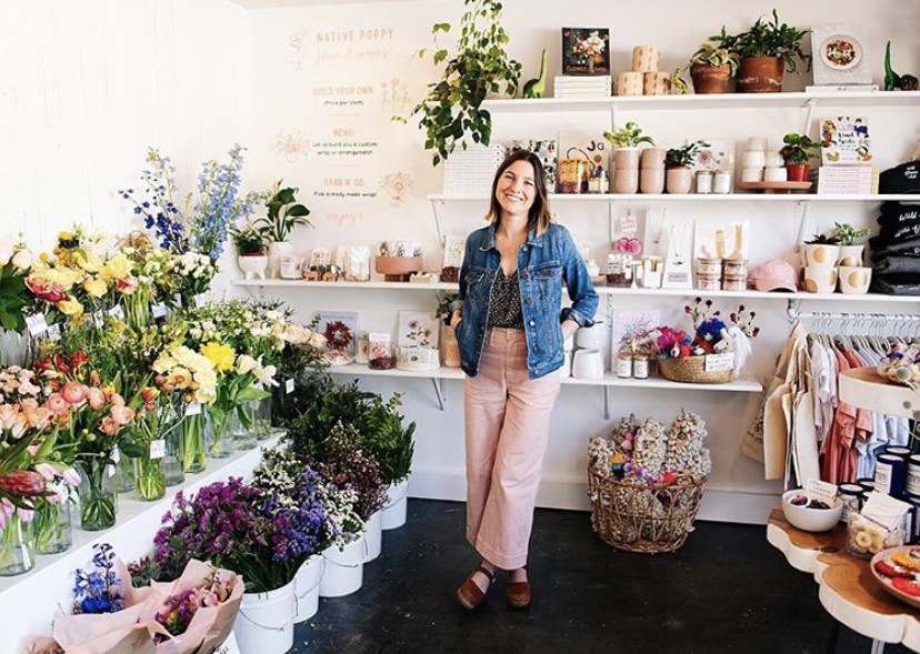 Natalie Gill stands in the center of a floral boutique wearing pink pants and a blue denim jacket