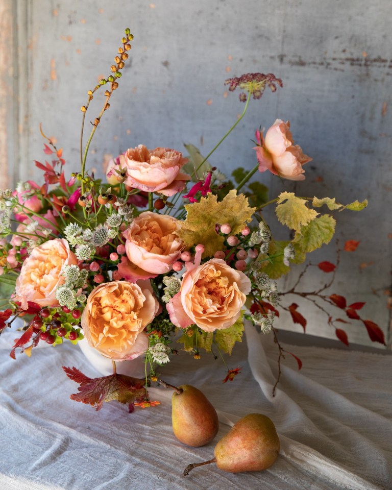 An orange and pink floral arrangement sits on a beige tablecloth next to two golden pears