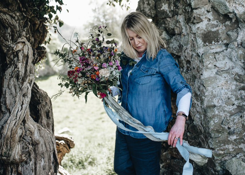 Janne Ford stands between two trees holding a floral arrangement tied with long, gray-blue fabric