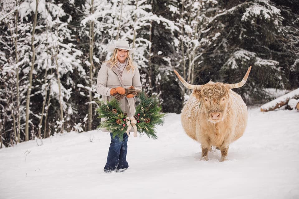 BB Podcast guest, Rachelle Willows, holds a festive wreath while standing in a snowy forest next to a cow 