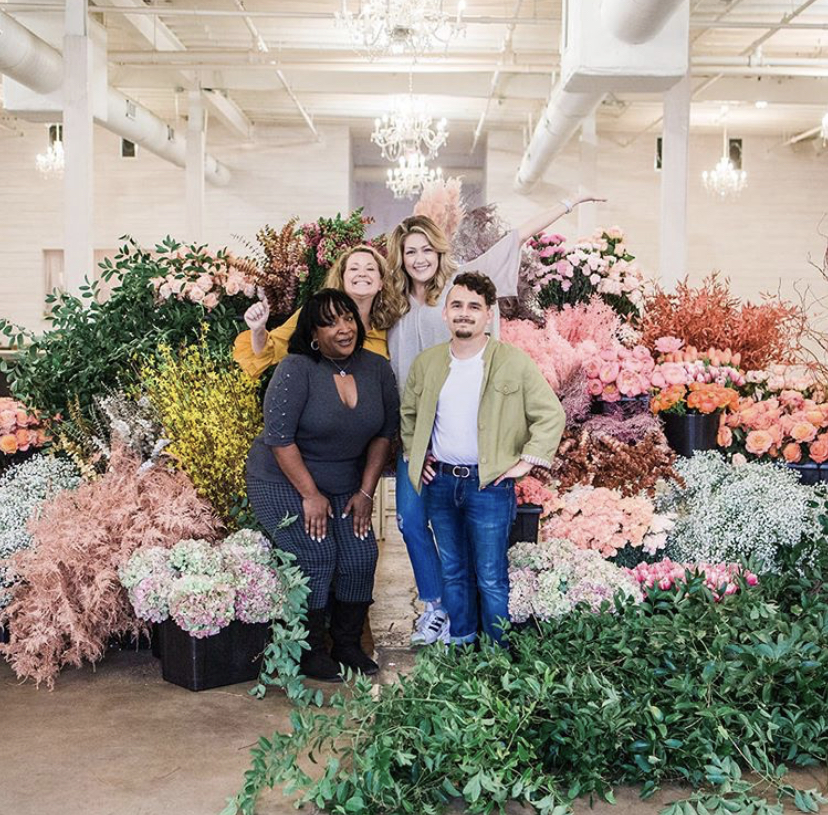A group of floral designers stand in a room full of various-colored buckets of flowers and arrangements 