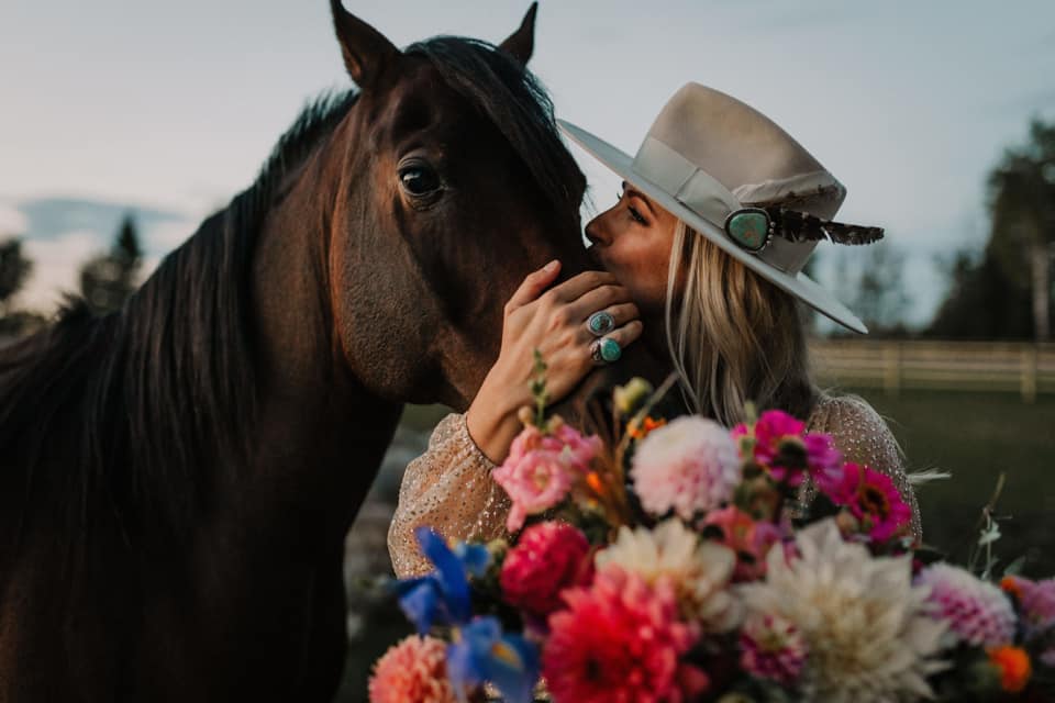 BB podcast guest, Rachelle Willows, holds a colorful floral arrangement while hugging a brown horse
