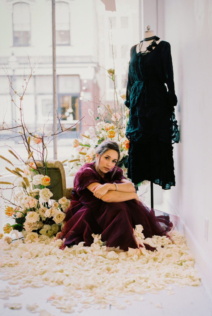 Model sitting on white rose petals next to a black display dress during a floral photoshoot