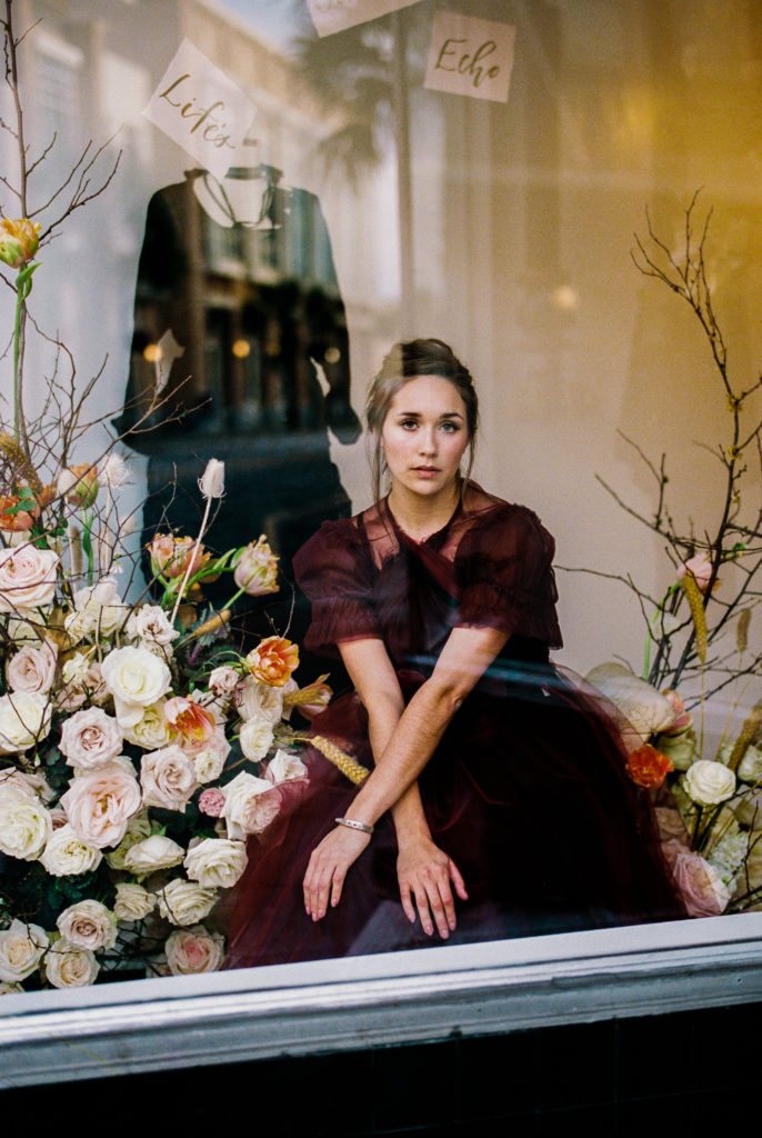 Model in clothing boutique window wearing a burgundy dress next to a white floral display