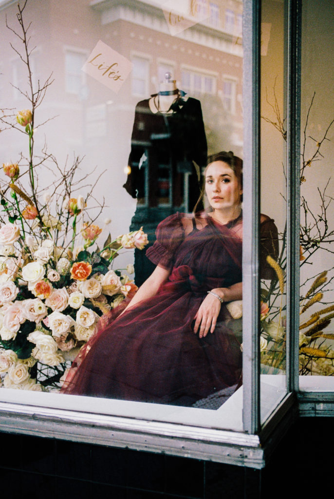 Model in burgundy dress in shop window with flower display by On a Limb