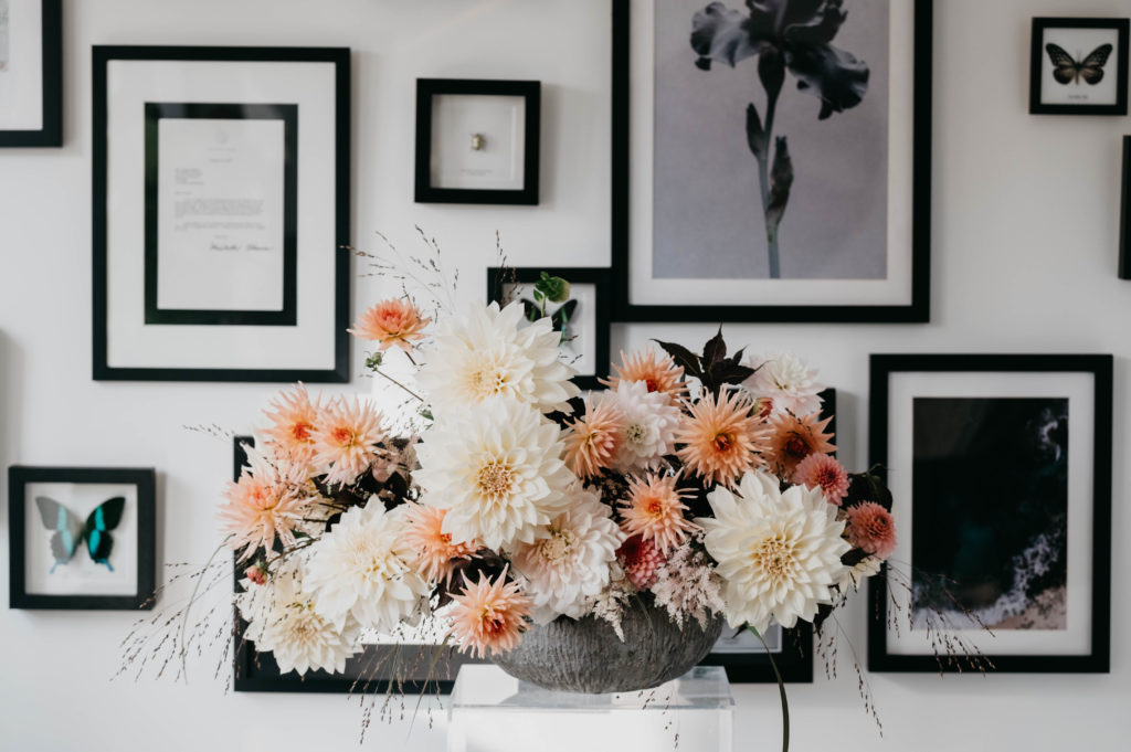 A floral arrangement with salmon Barberton daisies sitting in front of a white wall with monochrome art