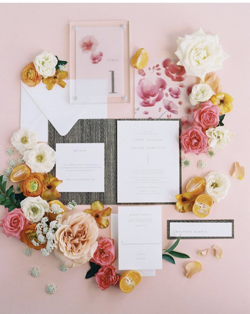 Flatlay consisting of a wedding invitation suite, an acrylic table number, and blooms in citrus colors