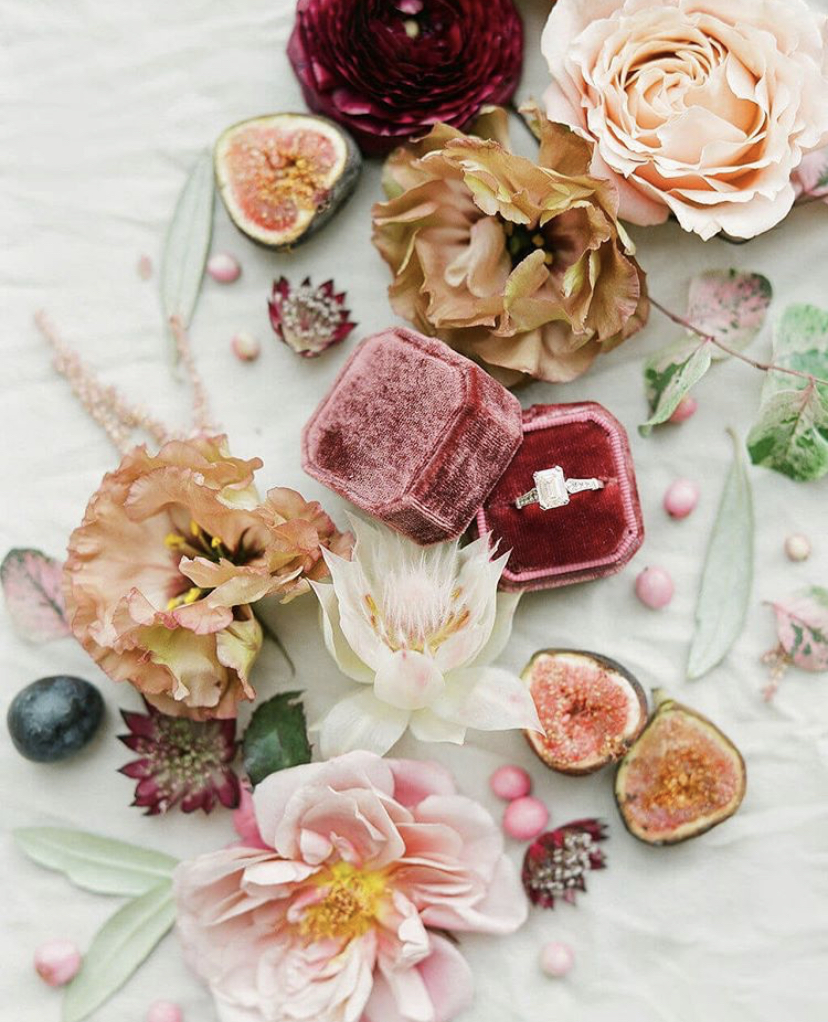 A diamond engagement ring in a burgundy velvet box featured in a styled flatlay of figs and flowers on a linen tablecloth