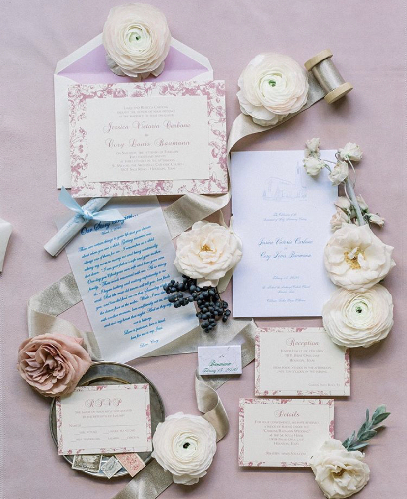 Flatlay of a wedding invitation, taupe ribbon on a wooden spool, white roses and ranunculus, and a silver plate holding vintage stamps on a lavender background