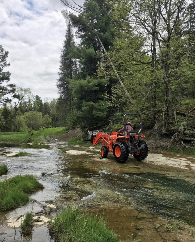 Dawson drives a tractor through the creek at Roam Flora flower farm to repair a fence