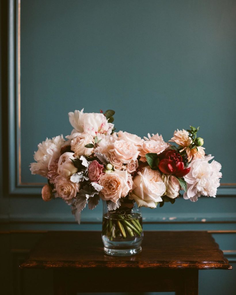 Peach neutral bouquet of roses and peonies designed by The Wild Mother sitting in a glass on water on a table 