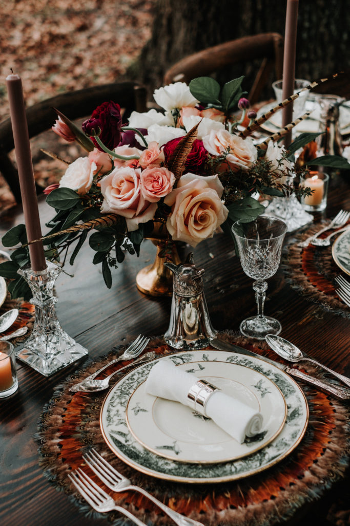 Styled shoot tablescape with autumn-colored centerpiece of flowers in a brass compote bowl next to a place setting of fine china on a feather charger
