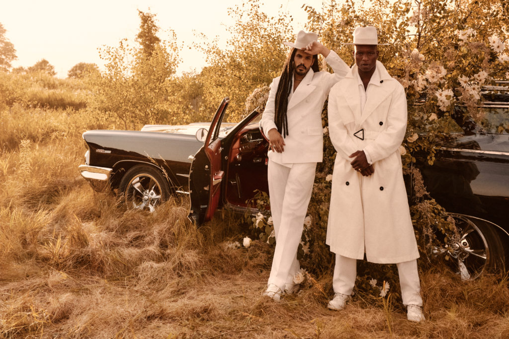  high fashion photo shoot in a field with two men standing in front of an abandoned car overgrown with vines and flowers