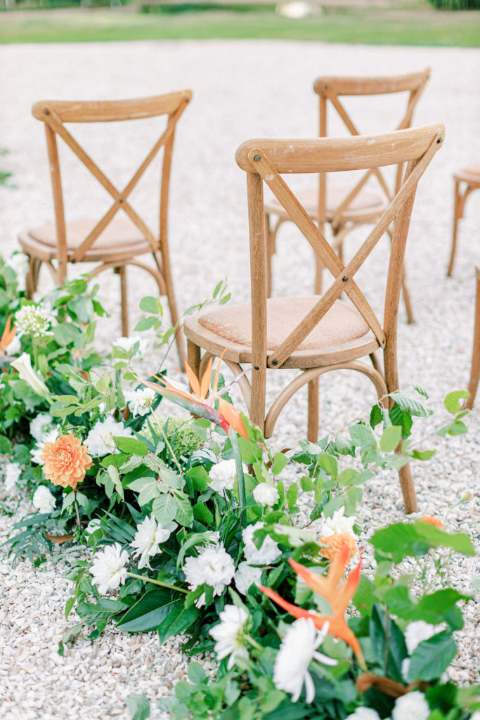 Outdoor wedding ceremony aisle lined with garland of orange and white flowers by Floraison Paris next to wooden chairs for guests