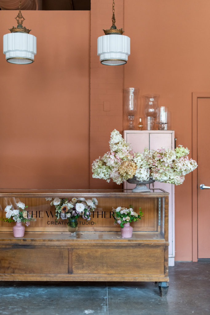 The antique wood and glass counter at The Wild Mother Studio sitting in front of a terra cotta colored wall with antique oendant lights hanging above and pastel flower arrangements sitting on the counter 