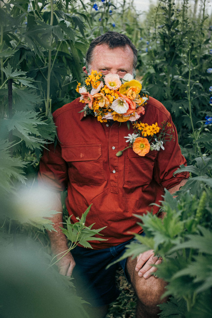Flowered fella kneeling between rows of delphinium while wearing a flower mask made of yellow and orange poppies