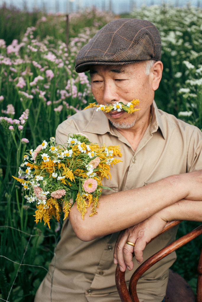 Flowered fella sitting in a chair inside a hot house on a flower farm while wearing a flower mustache and floral armband of yellow flowers