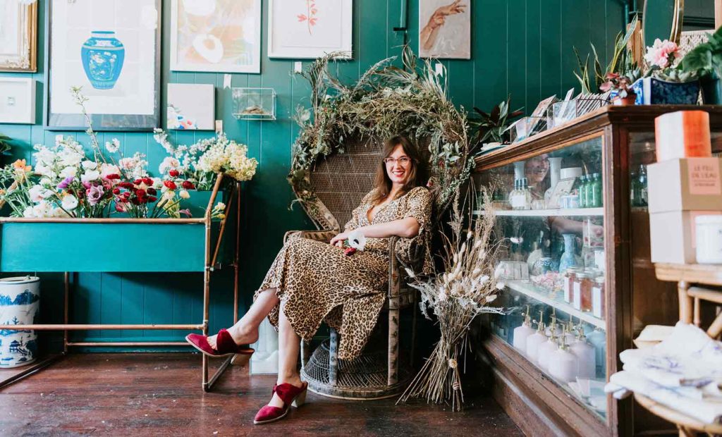 Holley Simmons, owner of the She Loves Me flower shop, sitting in a rattan chair surrounded by fresh and dried flower displays
