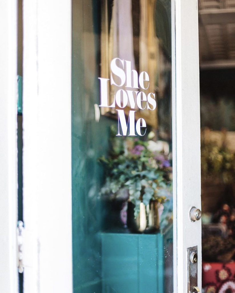 Clear glass door with the name of She Loves Me painted in white sits ajar inviting you into the flower shop