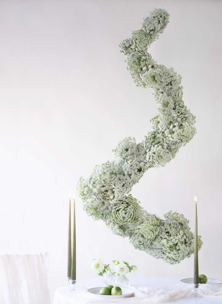 Hanging floral installations of Queen Anne's Lace forming a spiral above a white table with green taper candles and bud vases of white flowers