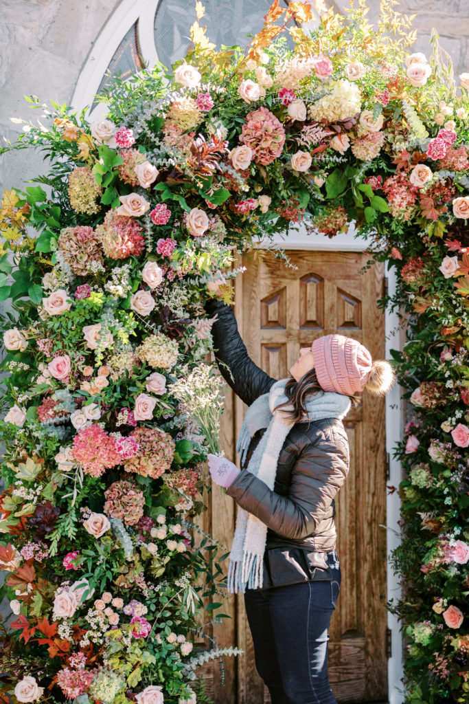 Janie Gerestein of Flowers by Janie places flowers in a foam free floral arch installation surrounding the chapel doors at a wedding