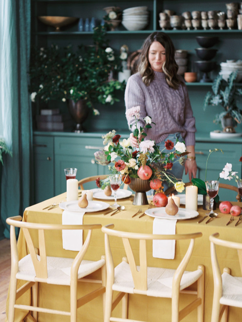 Floral designer Ginny Early standing behind a tablescape of flowers and fruit created in her Enemies of the Average studio