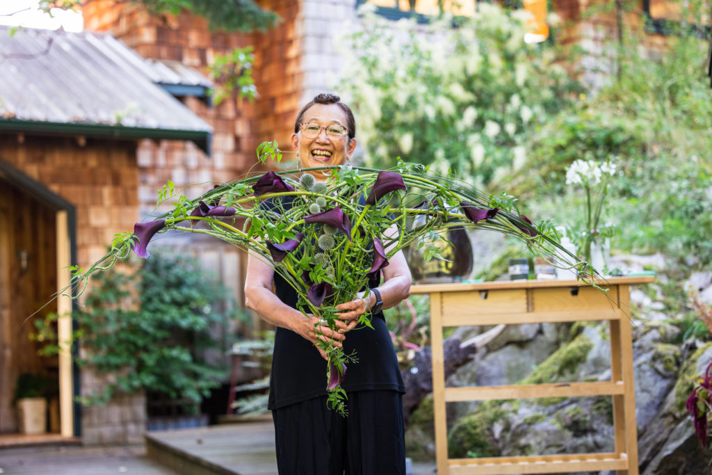 Freesia Summit co-founder Hitomi Gilliam AIFD in her personal garden holding a bouquet of eggplant colored callas she designed