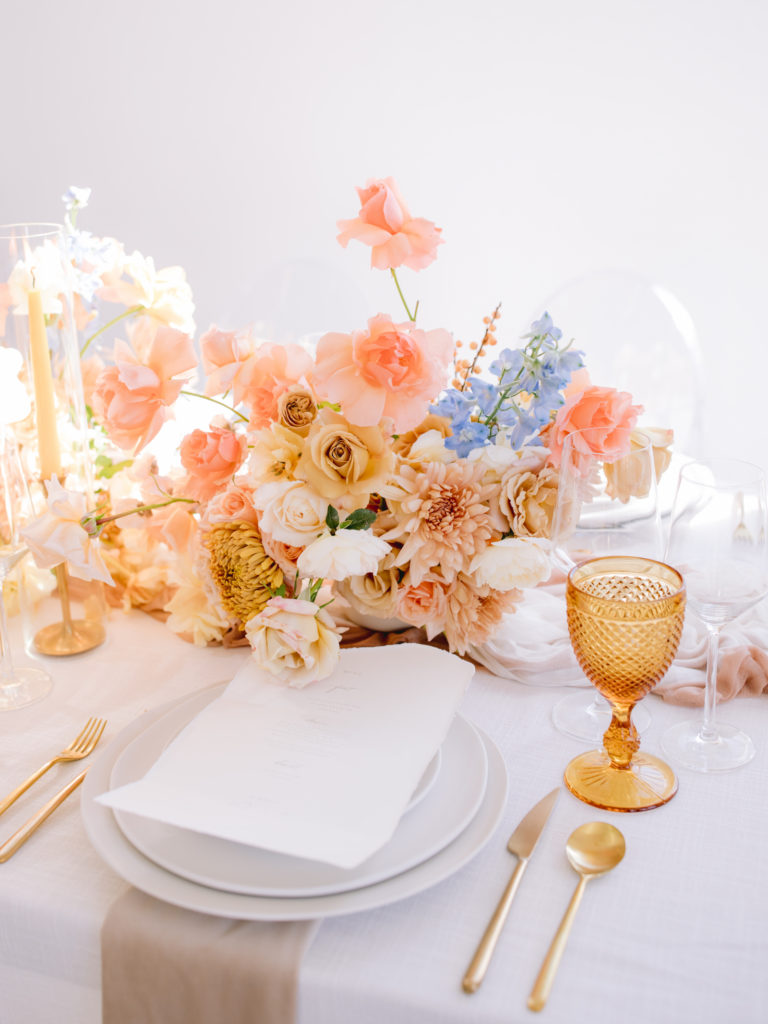 Golden yellow, peach, and blue flowers in a centerpiece by Le Bloomerie next to an amber glass goblet
