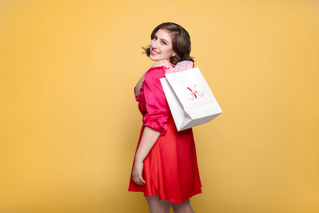 Storytelling strategist and brand communication expert Yasamin Salavatian dressed in a red and standing in front of a yellow background while holding a gift bag with her business logo on it