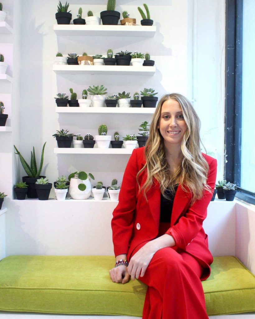 Dressed in a red suit, Popupflorist owner Kelsie Hayes sits on a window seat in her NYC flower shop surrounded by potted plants